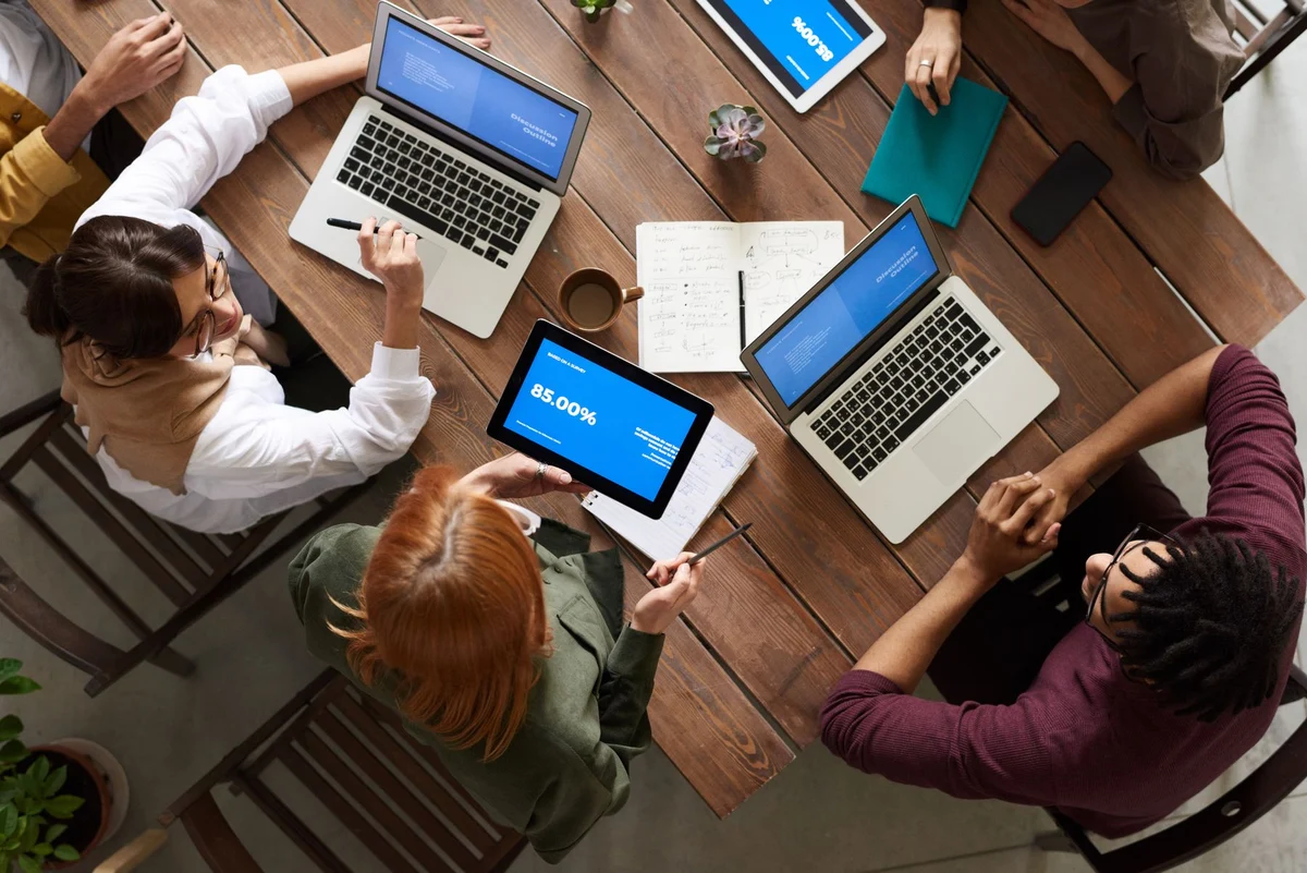 People reviewing information together using laptops, a tablet and notes at a meeting table.